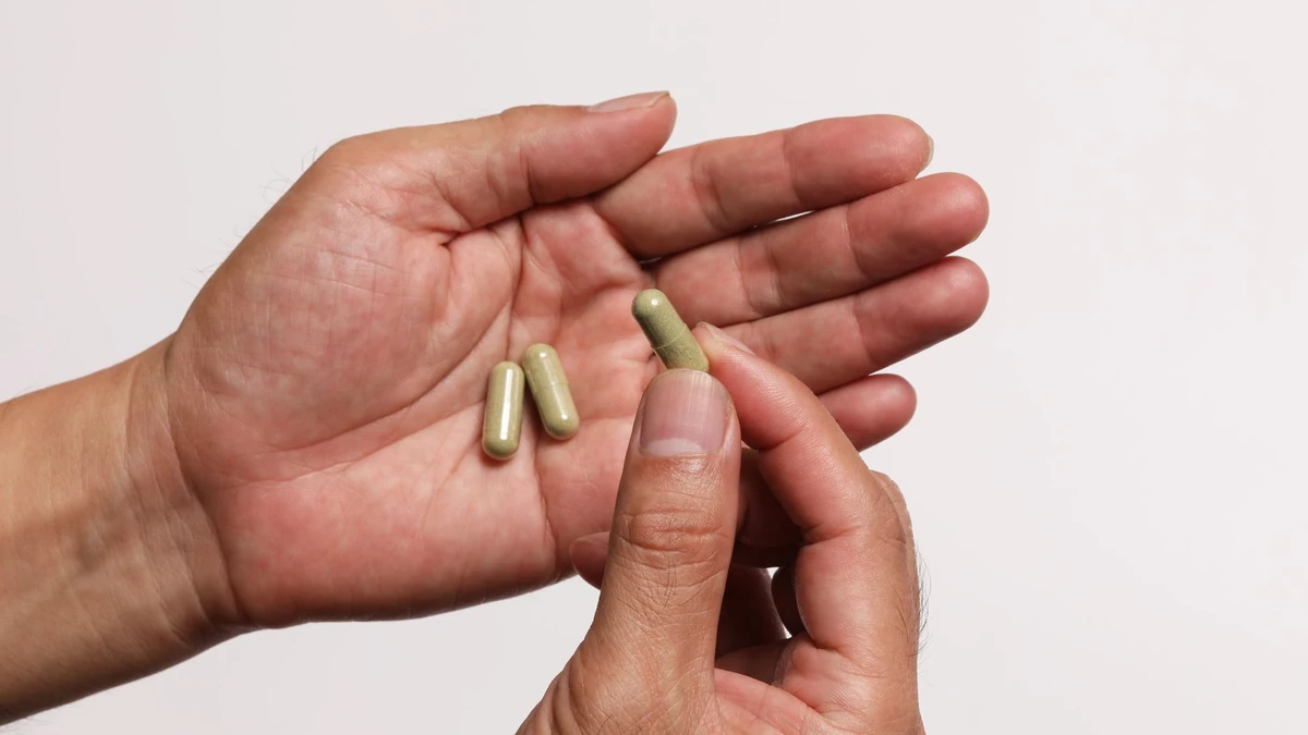 Mãos segurando cápsulas de ashwagandha, uma delas entre os dedos, sobre fundo claro